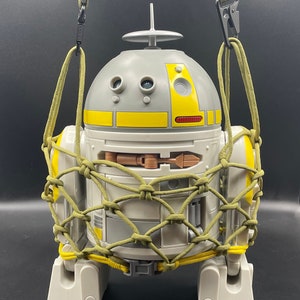May include: A white and gray robot with yellow accents is encased in a green and yellow net. The robot has a round head with a small antenna and three round eyes. The robot is standing on a black surface.