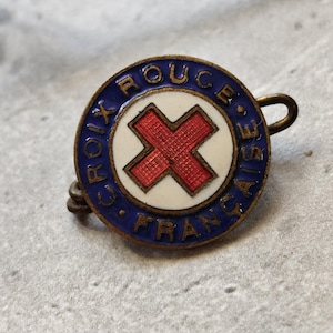 May include: A vintage enamel pin with a red cross on a white background, surrounded by a blue circle with the words "Croix Rouge Française" in gold lettering.