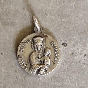 Pode incluir: Pingente prateado com imagem religiosa. O medalhão redondo mostra uma mulher coroada segurando uma criança, com a inscrição "SALVS POPVLI ROMANI". Possui uma pequena argola para uma corrente. Cor prateada.