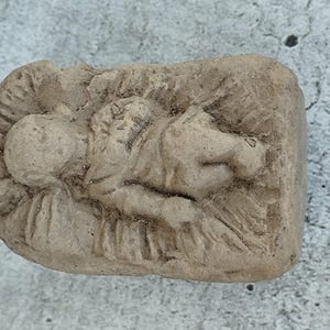 Può includere: Una statuetta in pietra beige testurizzata che raffigura una piccola figura distesa all'interno di una mangiatoia rettangolare. La superficie presenta una finitura opaca e usurata con dettagli intagliati che rappresentano la forma e la paglia.