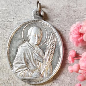 Peut inclure: Pendentif ovale argenté avec un relief détaillé d'une figure auréolée, tenant une palme. Le pendentif a une boucle en haut pour l'accrocher. Des fleurs roses sont en arrière-plan.