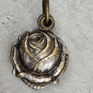 May include: A round metal pendant shaped like a blooming rose. The pendant features a dark, oxidized silver finish with highlights of polished gold on the edges of the petals. A small, circular metal loop is attached to the top for hanging.