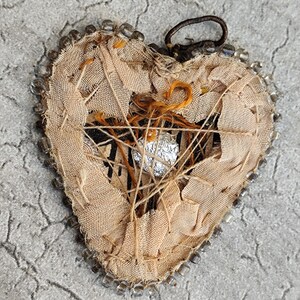 May include: A heart-shaped ornament crafted from frayed beige fabric with a dark brown loop at the top. The edges are lined with clear glass beads. The center features a web of tan thread, orange yarn, and a small, crumpled piece of silver foil.