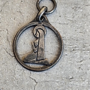 Peut inclure: Pendentif argenté représentant une personne agenouillée en prière dans un cadre circulaire. Le pendentif a une boucle en haut pour l'attache et un aspect vieilli et antique. Le fond est une surface grise texturée.