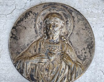 May include: A circular, antique metal religious medallion featuring a detailed relief of a figure with a halo, hands on chest, and a heart. The medallion has a weathered, aged appearance with shades of silver and bronze.