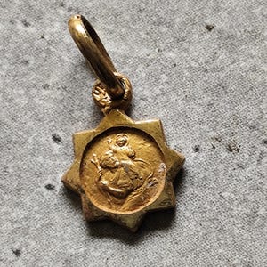 May include: A gold-colored pendant with a starburst frame. The pendant features a circular image of a religious figure carrying a child. The bail is attached to the top of the pendant.