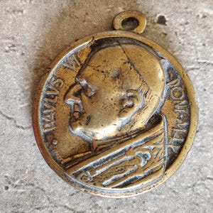 Pode incluir: Medalhão de bronze vintage com o perfil de um homem e a inscrição "PAVLVS VI PONT MAX". O pendente circular tem uma argola para pendurar e mostra sinais de idade e desgaste, com um design em relevo detalhado.