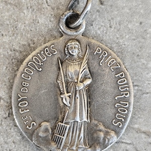 May include: A round silver-toned metal religious pendant featuring a relief of a figure holding a sword and a palm frond, with a grid-like object at their feet. The text "STE FOY DE CONQUES" and "PRIEZ POUR NOUS" is embossed along the circular edge.