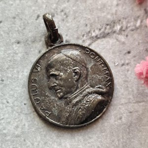 Pode incluir: Pingente de metal prateado com um retrato de perfil do Papa Paulo VI. O pingente tem a inscrição "PAULUS VI PONT. MAX." ao redor da borda.