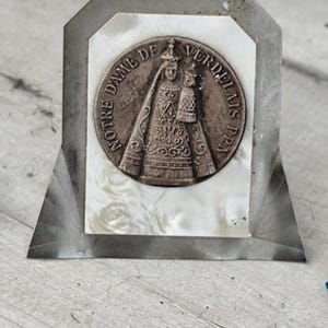 Puede incluir: Un icono religioso vintage con un medallón de bronce con la inscripción "NOTRE DAME DE VERDELAIS PRN." El medallón representa una figura vestida con una túnica que sostiene a un niño, sobre un fondo de nácar dentro de un marco plateado. El marco tiene una base triangular.