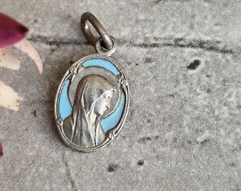 Peut inclure: Pendentif ovale en argent avec une bordure bleu clair et un profil d'une figure religieuse. Le pendentif a une boucle en haut pour une chaîne. Le fond est une surface grise texturée.