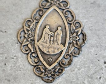 May include: Silver-toned metal pendant with an oval central design depicting two figures. The pendant is framed by an ornate filigree border with floral and scroll details. A small cross is at the bottom of the oval.