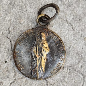 May include: A circular bronze-toned metal pendant featuring a raised relief of a figure holding a staff. The text "SAINTE ADELE PRIEZ POUR NOUS" is embossed around the perimeter. The pendant includes a dark metal jump ring at the top.