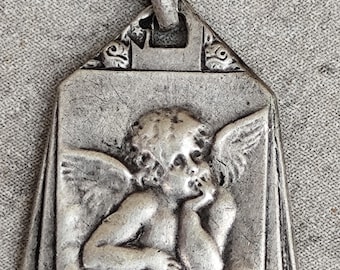 May include: A silver-toned metal pendant featuring a relief of a cherub with wings, resting its chin on a hand. The rectangular pendant has a tapered top, a bail for hanging, and decorative floral motifs at the bottom corners.