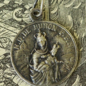 May include: Silver pendant with a depiction of the Virgin Mary holding a baby Jesus. The pendant has the words "Madre de Buron" inscribed around the edge.