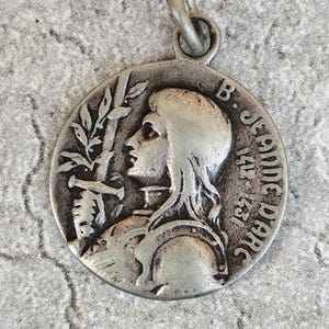 May include: A circular silver-toned metal pendant featuring a relief profile of a figure in armor holding a sword. The text B. JEANNE DARC 1412-1431 is embossed on the right side. The pendant includes a small circular bail at the top for attachment.