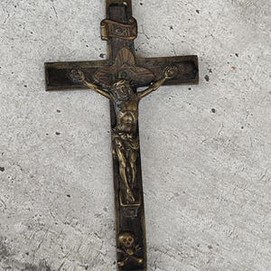 May include: A bronze-toned metal crucifix featuring a central figure of Jesus on a dark cross. Above the figure is a small scroll with the letters INRI, and at the base of the cross, there is a skull and crossbones emblem. A small loop is at the top.
