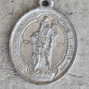 Op de afbeelding: Vintage zilverkleurige Sint-Christoffelmedaille. De ovale hanger heeft een reliëfbeeld van Sint-Christoffel die een kind draagt, omringd door tekst. De medaille heeft een lus aan de bovenkant om op te hangen.