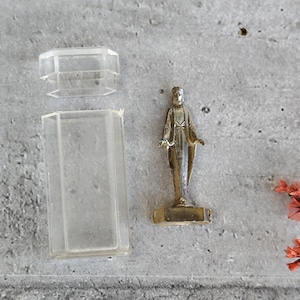 May include: A small, antique-style statue of a figure with outstretched arms, standing on a rectangular base. The statue is next to a clear, rectangular plastic box with a lid. The background is a textured grey surface.