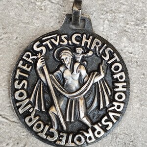 May include: A circular silver-toned metal pendant featuring a relief of Saint Christopher carrying a child on his shoulders while holding a staff. The border is inscribed with the Latin text "SANCTUS CHRISTOPHORUS PROTECTOR NOSTER" in raised lettering.