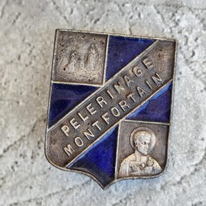 Puede incluir: Un broche vintage plateado en forma de escudo con detalles de esmalte azul. El broche presenta el texto "PELERINAGE MONTFORTAIN" e imágenes pequeñas. El diseño incluye una figura religiosa y un paisaje.