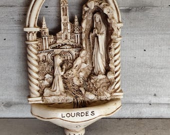 Könnte beinhalten: Beige religiöses Wandrelief mit detaillierter Darstellung einer Kirche, Figuren und dem Wort "LOURDES". Das Bogendesign umfasst ein Becken am Boden mit dem Wort "PAX". Das Gesamtthema ist religiös.