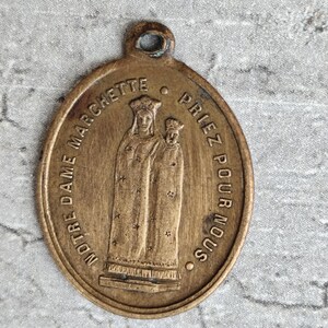 Peut inclure: Pendentif ovale en bronze vintage avec une image en relief de deux personnages, probablement religieux. Le pendentif porte l'inscription "NOTRE DAME MARCHETTE PRIEZ POUR NOUS". Il possède une boucle en haut pour l'accrocher.