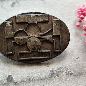 Puede incluir: Un pin de metal plateado con un diseño geométrico que presenta un trébol de cuatro hojas en el centro.