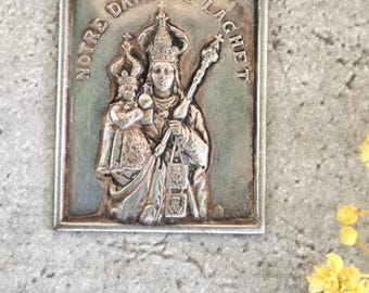 May include: A rectangular silver-toned religious pendant featuring an embossed image of a crowned woman holding a staff and a child. The text "NOTRE DAME DE LACHET" is inscribed around the image. The pendant has a detailed, vintage appearance.