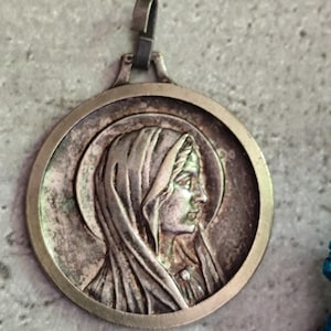 May include: A circular, silver-coloured pendant depicting a woman in a veil, set within a round frame. The pendant has a loop at the top for hanging and shows signs of wear, possibly vintage or antique.