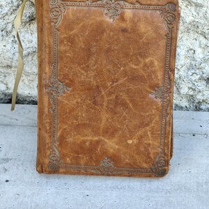 Puede incluir: Un libro antiguo, encuadernado en cuero, con un aspecto desgastado y texturizado. La cubierta es de color marrón cálido con diseños decorativos en relieve en las esquinas. Una cinta de color crema sobresale de la parte superior. La apariencia envejecida del libro sugiere que es una antigüedad o un artículo artesanal.