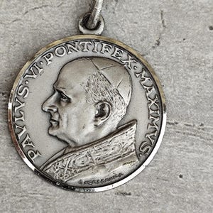 Pode incluir: Um pingente redondo prateado com um retrato de perfil de um homem, rodeado por texto. O pingente tem uma argola na parte superior para pendurar. O texto ao redor da borda diz "PAULUS VI PONTIFEX MAXIMUS".