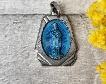 Puede incluir: Colgante plateado con un óvalo de esmalte azul que representa una figura religiosa y un niño. El colgante tiene un marco geométrico decorativo y un lazo para una cadena. Flores amarillas en el fondo.