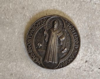 May include: A round, antique-style metal medallion with an embossed image of a robed figure. The figure is surrounded by text and other symbols. The medallion has a dark, aged appearance, suggesting it is vintage or a replica of an older piece.