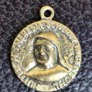 Puede incluir: Un colgante redondo de color oro antiguo con un retrato de una mujer con un tocado. El colgante tiene el texto "MUTTER THERESIA SCHERER" alrededor del borde y un pequeño lazo en la parte superior para una cadena.