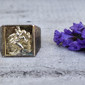 May include: A vintage silver ring with a rectangular face featuring a detailed relief of a figure. The figure is in a gold tone, set against a darker background. A cluster of purple flowers is in the background.