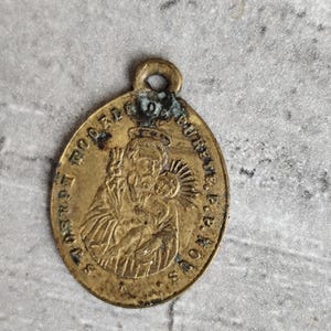 Può includere: Ciondolo ovale dorato antico con un anello nella parte superiore. Il ciondolo presenta un'immagine in rilievo di una figura religiosa che tiene in braccio un bambino, circondata da testo. Il ciondolo ha un aspetto usurato e invecchiato.