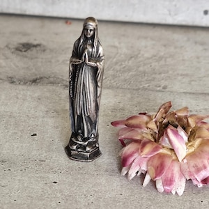 Könnte beinhalten: Kleine, silberfarbene Statue einer Frau mit gefalteten Händen, die ein drapiertes Gewand trägt. Die Statue steht neben einer getrockneten, rosa-cremefarbenen Blume auf einer grauen Oberfläche.