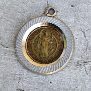 Puede incluir: Colgante redondo de estilo antiguo con una imagen religiosa detallada. El medallón central presenta una figura con una túnica, rodeada de texto. El colgante tiene un centro dorado y un anillo exterior plateado y estriado. Un pequeño lazo en la parte superior para una cadena.