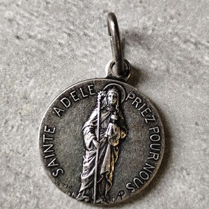 Puede incluir: Colgante plateado con la imagen de una santa sosteniendo un báculo y un niño. El colgante tiene la inscripción "SAINTE ADELE PRIEZ POUR NOUS" alrededor del borde. Un pequeño lazo en la parte superior para colgar.
