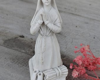 May include: A white statue of a kneeling figure with hands clasped in prayer. The figure wears a veil and a long robe. A rosary hangs from the neck. At the base are two small shoes and a bundle. Pink dried flowers are in the background.