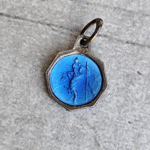 Puede incluir: Colgante vintage con un centro de esmalte azul que representa una figura que lleva un bastón. El colgante tiene un marco de metal plateado y un pequeño lazo en la parte superior para una cadena. El colgante es de forma octogonal.