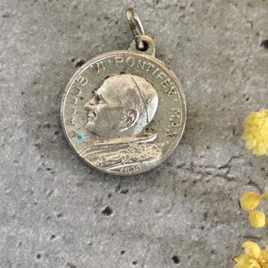 Pode incluir: Pingente redondo prateado com um retrato de perfil e texto. O pingente tem uma argola para uma corrente. O fundo é uma superfície cinza texturizada. O pingente é provavelmente de metal.