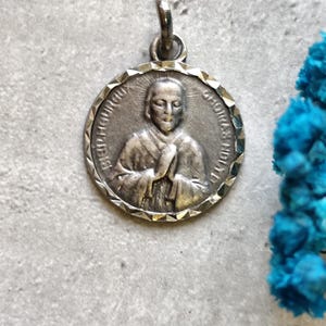 May include: Silver pendant featuring a religious image of a person with hands clasped in prayer. The pendant has a scalloped edge and the words "BEATUS IN DOMIN" and "SANCTUS IN DIE" are inscribed around the image.