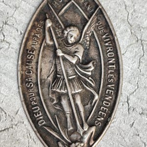 May include: An oval-shaped silver-toned metal medallion featuring a relief of an angel with wings holding a spear and standing over a figure. The border contains the embossed French text: DIEU POUR SA CAUSE AURA DES HOMMES ET TANT QUE VIVRONT LES VENDEENS.