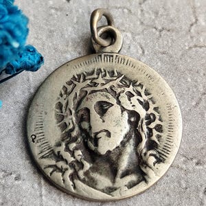 Könnte beinhalten: Silberner Anhänger mit einem detaillierten Relief eines Männergesichts mit Dornenkrone. Der Anhänger hat oben eine Öse zum Anbringen an einer Kette.