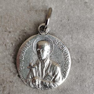 Peut inclure: Un pendentif argenté représentant en relief une personne tenant une croix, avec l'inscription "S. JOANNE BERGMANNS OP.F.A." sur le bord. Il comporte une petite boucle en haut pour une chaîne.