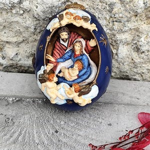 Può includere: Un ornamento blu a forma di uovo con una scena della Natività. La scena include Maria, Giuseppe e il bambino Gesù, circondati da cherubini. L'ornamento ha una parte superiore color oro ed è ambientato su uno sfondo strutturato.