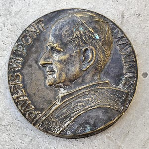 Pode incluir: Medalha redonda de bronze antigo com o perfil de um homem usando um chapéu. A medalha tem letras em relevo ao redor da borda. O bronze tem uma aparência desgastada com áreas de oxidação verde.