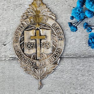 May include: A silver and gold metal pin with a cross and open book design. The pin reads "Enseignement Libre Diocese de Lucon 1831 1931".
